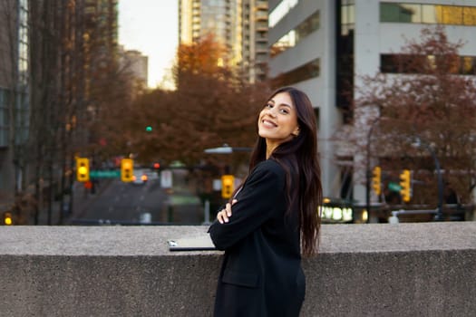 Smiling young businesswoman standing in front of a modern building with autumn foliage.