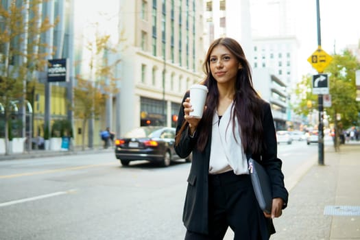 Confident young latin businesswoman holding a coffee cup walking down a city street.