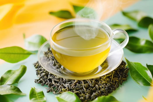 Steaming cup of green tea is standing on a wooden table with fresh tea leaves scattered around it