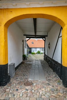 A picturesque cobblestone alley framed by a bright yellow archway, leading to a charming white house with a red roof. The setting conveys a sense of history and tranquility in ebeltoft in denmark
