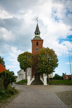 ebeltoft denmark,A striking brick church tower stands majestically under a dramatic cloudy sky. The surrounding landscape features lush trees and traditional architecture, creating a serene and timeless ambience