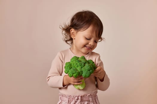 adorable child girl holding fresh green raw broccoli on beige background. healthy baby food. kid loves vegetables