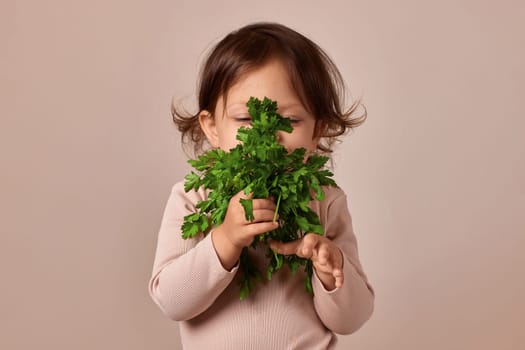 little child girl holding fresh green parsley on beige background. healthy baby food.