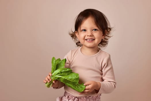 smiling child girl holding fresh green salad on beige background. healthy baby food.