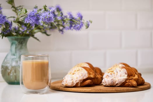 Delicious fresh croissants on wooden tray and glass of coffee on white table, closeup, tasty breakfast