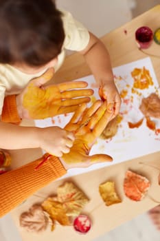 little child girl and mother painting hands and autumn leaves together. mom and baby showing hands in yellow paint. top view
