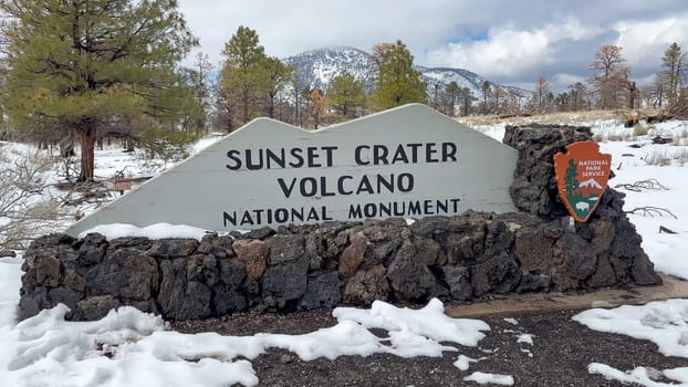 Flagstaff, Arizona, USA - 03.19.2024 - Sunset Crater Volcano National Monument entrance sign on a snowy winter day. The sign is made of wood and stone, and it is surrounded by snow-covered ground.