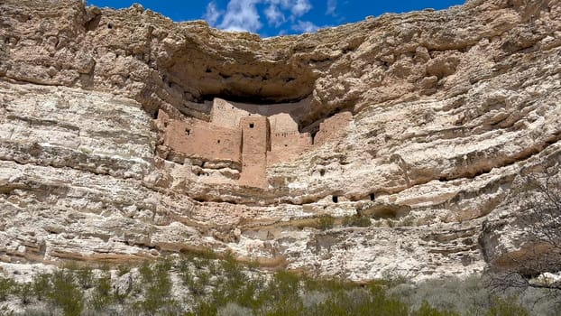 A low-angle view of Montezuma Castle National Monument in Arizona. The ancient Sinagua cliff dwellings are built high up into a limestone alcove. White clouds dot the blue sky above - USA