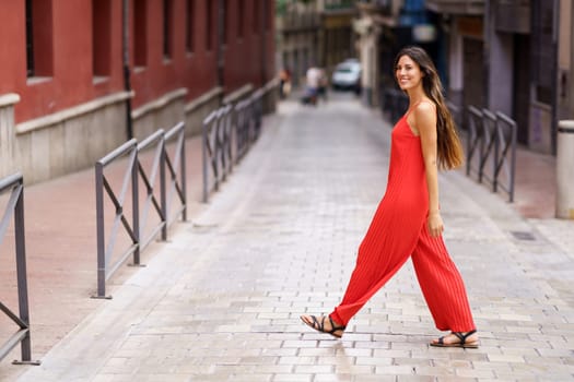 A stylish woman adorned in a vibrant red jumpsuit saunters gracefully along a scenic urban street, effortlessly showcasing her confidence and impeccable fashion sense for all to admire and appreciate