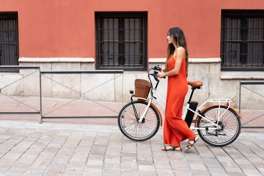 A fashionable woman in a vibrant red dress strolls beside her chic ebike along a colorful wall, embodying an elegant urban lifestyle that showcases her unique style and charisma