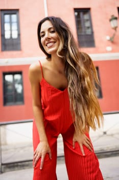 A fashionable woman, showcasing her long, flowing hair, strikes a confident pose in a strikingly vibrant red outfit, set against a charming urban backdrop that enhances her style and mood