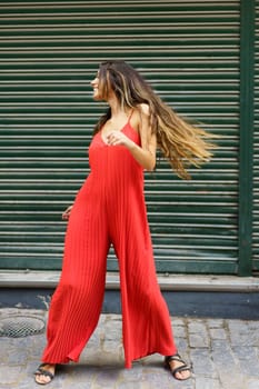 A glamorous young woman exudes confidence as she poses playfully in a trendy red jumpsuit, beautifully showcasing modern fashion against a vibrant green backdrop that complements her stylish look