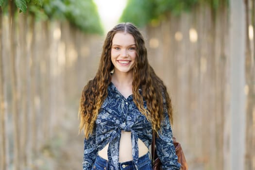 A cheerful young woman with curly hair stands confidently at a charming pathway, which is beautifully lined with an array of vibrant trees, capturing a truly magical moment of natural beauty