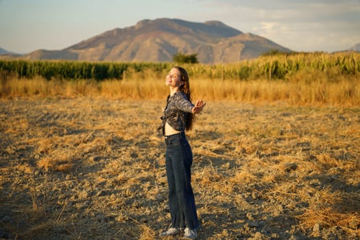 A carefree woman stands joyfully in a vast golden field, her arms wide open as she embraces the stunning beauty of the majestic mountains, which are illuminated by the warm, golden rays of the sunset