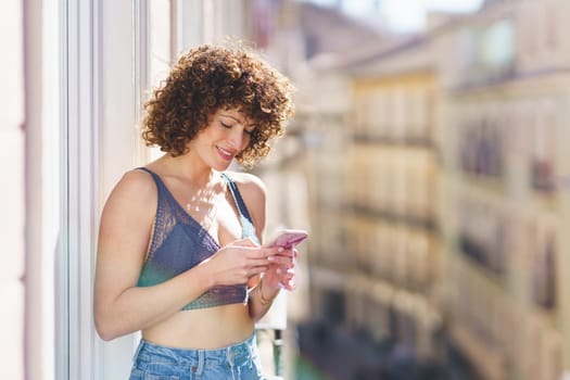 Side view of positive young female in bra and jeans with curly hair smiling while browsing smartphone on sunny balcony