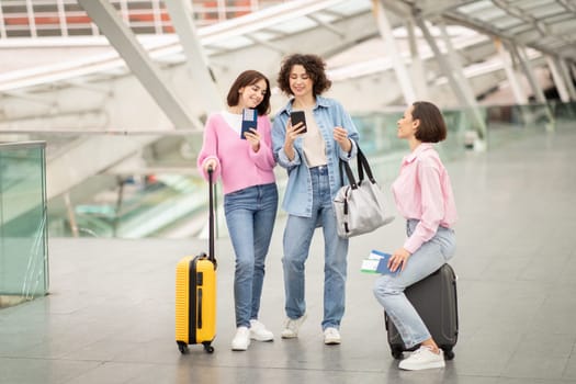 Three friends are gathered at a contemporary travel terminal, enjoying their time while checking travel plans on their smartphones. They stand with luggage, engaged in conversation.
