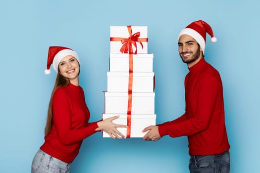A happy couple wearing red sweaters and Santa hats joyfully holds a stack of decorated Christmas gifts. The blue backdrop enhances the festive spirit.