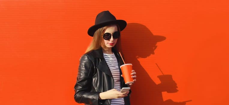 Stylish modern young woman with phone, girl using smartphone holding cup of coffee in black hat, leather jacket on red wall background with shadows