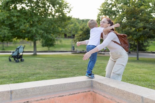 A woman is holding a child in her arms. The child is wearing a white shirt and blue jeans. The scene takes place in a park, with a bench and a stroller nearby