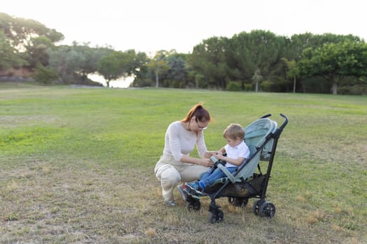 Mother is fastening the seat belt on a stroller for her son while enjoying a beautiful day at the park