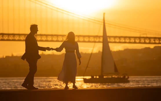 Silhouette of a couple holding hands and dancing by a river with a bridge and sailboat in the background at sunset
