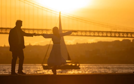 Silhouetted couple dancing by the river at sunset with a bridge and sailboat in the background