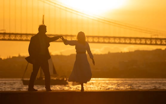 Silhouette of a couple holding hands and dancing on a pier by the river at sunset with a bridge in the background