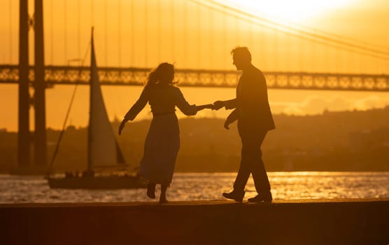 Silhouette of a couple holding hands and dancing by the river with a bridge and sunset in the background