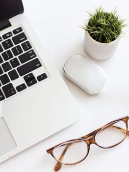 A clean, white desktop workspace featuring a laptop, wireless mouse, eyeglasses, and a small potted plant, embodying modern minimalist office design with copy space.