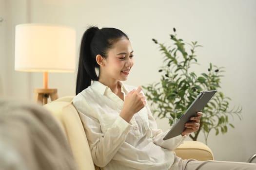 Smiling young woman wearing casual dressed using digital tablet on sofa at home.
