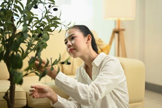 Young woman examining a potted plant in a living room. Home gardening and mindfulness concept.
