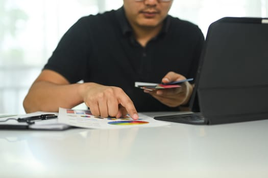 Young creative man working on a design project reviewing color chart and using laptop at desk.