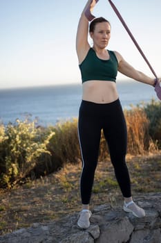 Woman is doing a workout with a resistance band on a clifftop with the ocean in the background