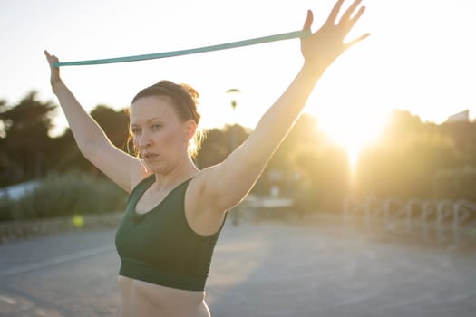 Athletic woman is using a resistance band while working out outside at sunset