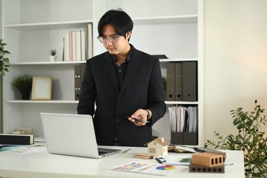 Young adult man using laptop, working on a design or architectural project at office.