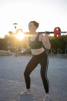 Woman is performing squats while lifting a weighted barbell outdoors during golden hour