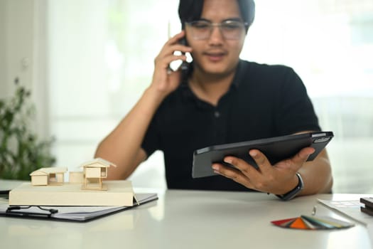 Architect man talking on cell phone and using digital tablet at creative workplace.