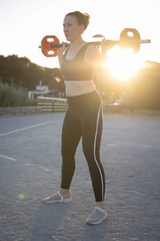 Young woman is performing squats with a weight bar outdoors at sunset