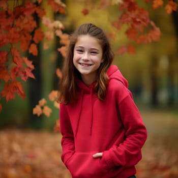 A young girl in a red hoodie smiles brightly in an autumn park surrounded by vibrant fall foliage.