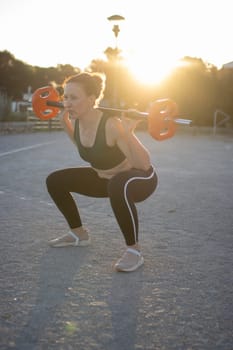 Woman is doing squats with a weight bar on her shoulders while exercising outdoor at sunset