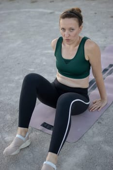 Woman in sportswear is taking a break from exercising, sitting on a yoga mat in an outdoor workout area