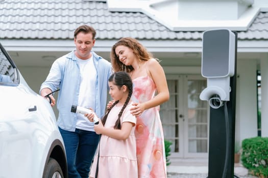 Happy little young girl learn about eco-friendly and energy sustainability as she help her family recharge electric vehicle from home EV charging station. EV car and modern family concept. Synchronos