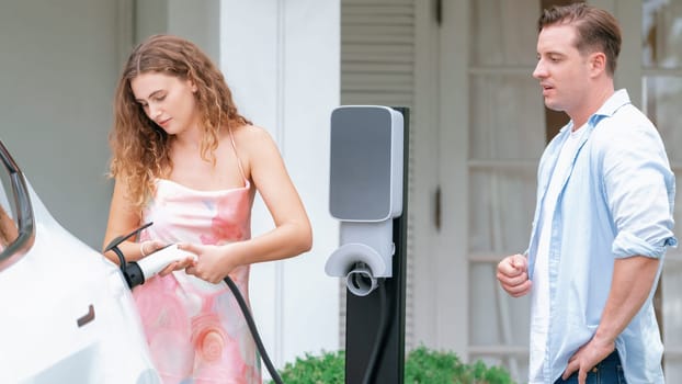 Happy and lovely couple with eco-friendly conscious recharging electric vehicle from EV home charging station. EV car technology utilized for residential home to future sustainability. Synchronos