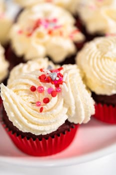 Freshly baked velvet cupcakes with white chocolate ganache frosting decorated with sprinkles.