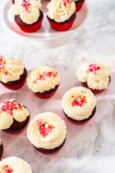 Freshly baked velvet cupcakes with white chocolate ganache frosting decorated with sprinkles.