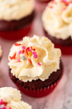 Piping white chocolate ganache frosting on top of red velvet cupcakes and topping with sprinkles.