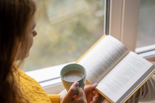 Young beautiful woman sitting by the window yellow knitted sweater read book, daily planner, notepad. Relax concept. Hold cappuccino glass of coffee with white foam. Text is out of focus