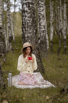 A young beautiful woman in a dress and a round hat reads a book outdoors in the forest and drinks tea. Romantic and vintage photo of a beautiful girl. Reading and relaxation