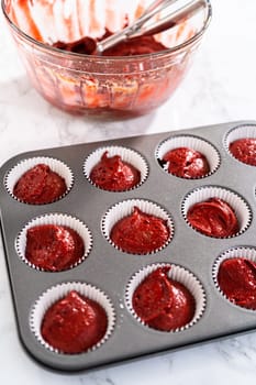 Scooping cupcake dough with dough scoop into cupcake pan lined with foil liners to bake red velvet cupcakes with white chocolate ganache frosting.