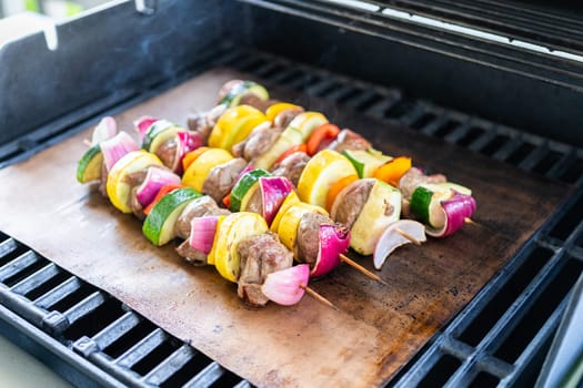 Skewered beef and fresh veggies sizzle on a copper grill mat over a gas outdoor grill.